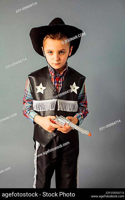 The little boy in a sheriff's costume is standing at the gray background. The boy is holding a toy gun