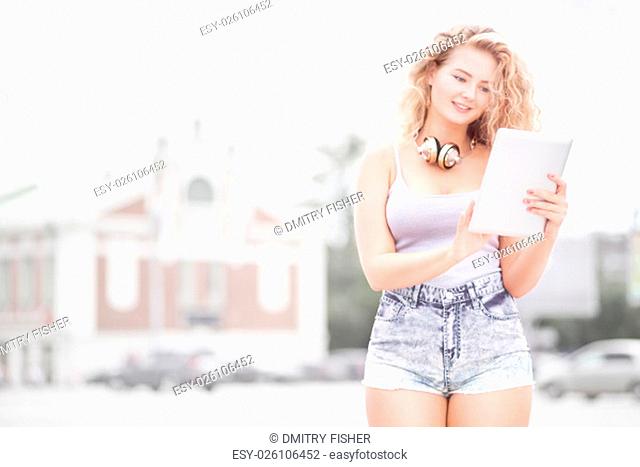Happy young woman with vintage music headphones around her neck, surfing internet on a tablet pc and posing against urban city background