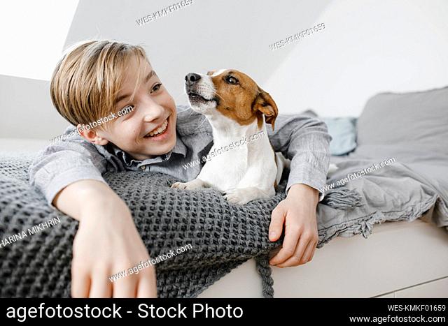 Smiling boy with arm around dog lying on bed at home