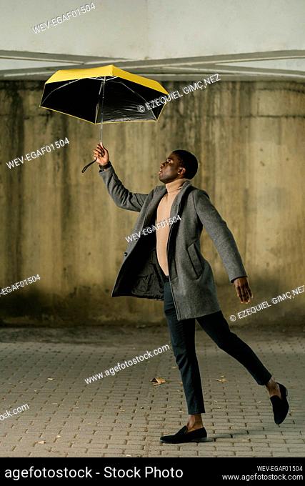 Young man wearing jacket holding umbrella while walking on footpath