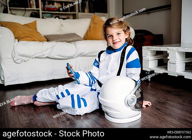 Smiling girl wearing space costume holding mobile phone sitting with helmet on floor at home