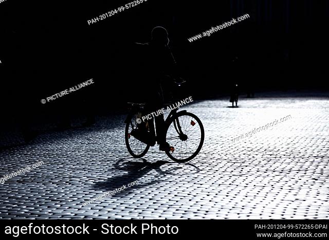 04 December 2020, North Rhine-Westphalia, Münster: A cyclist with a mouth and nose protector rides across the Prinzipalmarkt in strong backlighting