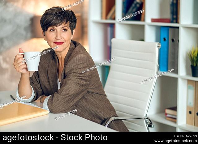 Morning in the office. Good-looking dark-haired woman sitting at the table with a coffee mug in hands