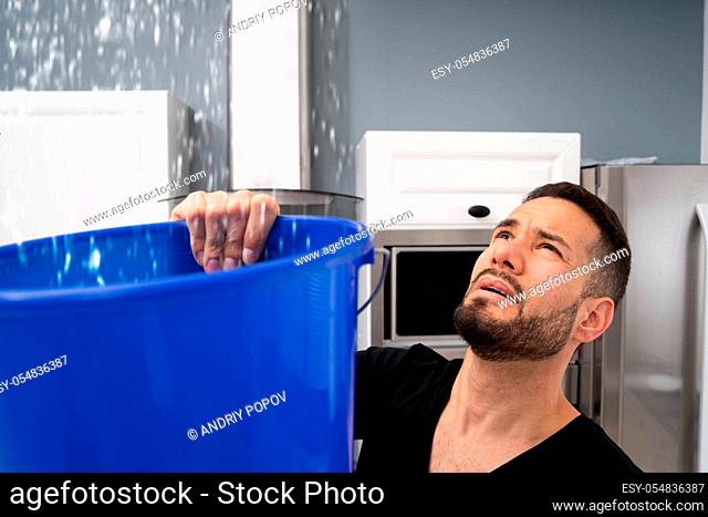 Worried Man Holding Bucket While Water Droplets Leak From Ceiling In Kitchen