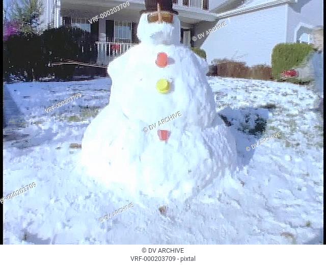 Children stand next to a snowman in front of their house