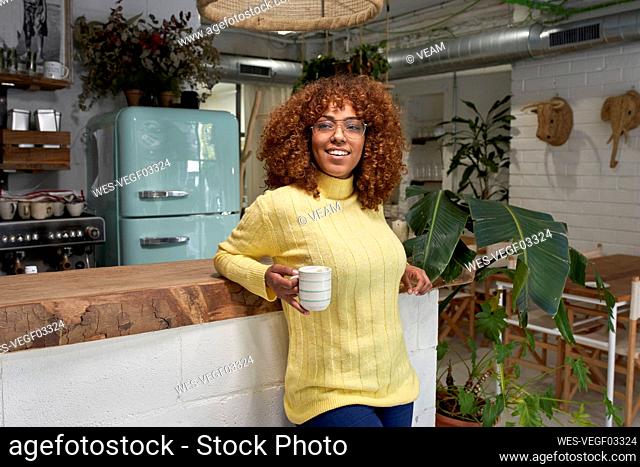 Smiling woman holding coffee cup while leaning on counter at cafe