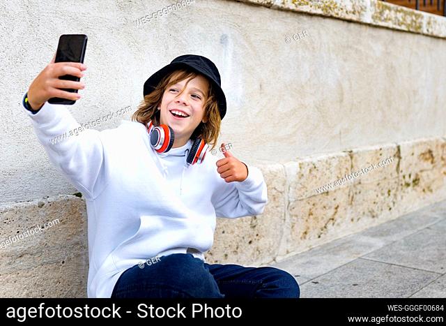 Boy wearing hat and headphones using mobile phone while sitting against wall