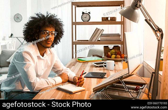 Creative man writes in Note Pad working laptop at Home work place. Arabic business or student in eyeglasses looking at camera. Freelance concept