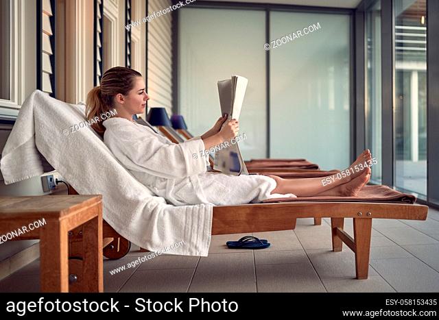 Young woman relaxing at a spa after a pampering treatment sitting on a day bed on a patio reading a newspaper in a white bathrobe