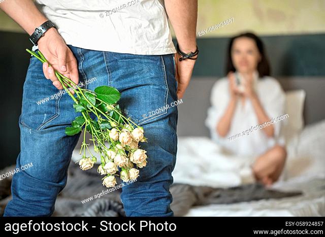 Guy in blue jeans and white T-shirt holds a bouquet of roses behind her back. He stands opposite his girl in a white shirt who sits on the bed