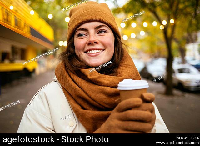 Smiling woman with disposable coffee cup looking away on sidewalk during autumn