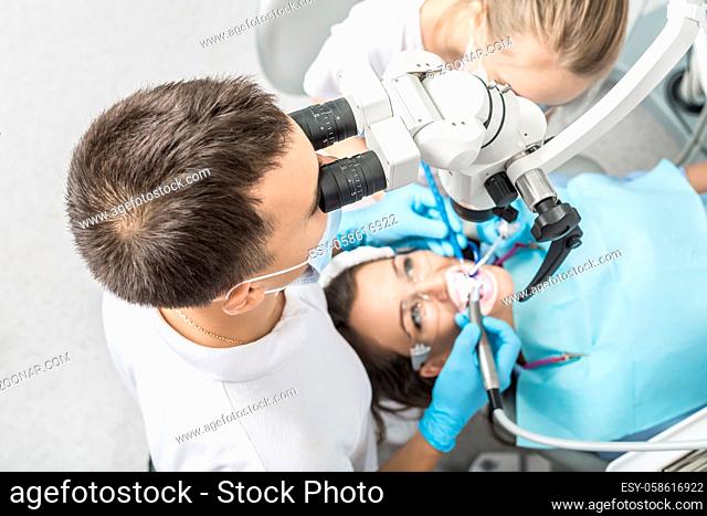 Young dentist looks at patient teeth using a dental microscope and holds dental instruments near her mouth. Female assistant helps the doctor