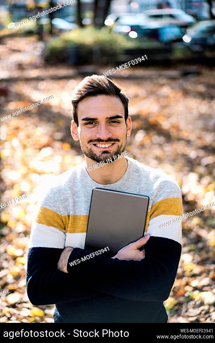 Happy handsome man holding digital tablet while standing with arms crossed at park