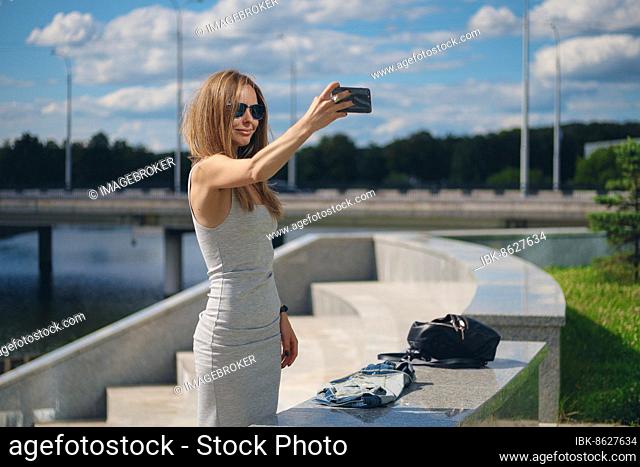 Attractive tourist girl making selfie with river bank on background in city park