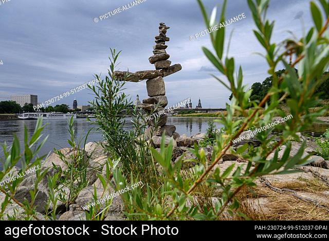 24 July 2023, Saxony, Dresden: A sculpture with piled up stones stands on the dried up bank of the Elbe below the Albert Bridge in front of the old town scenery...