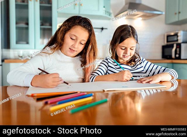 Sisters coloring with colored pencil on paper while sitting at dining table in kitchen