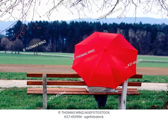Person with red umbrella sitting on bench under tree. Germany