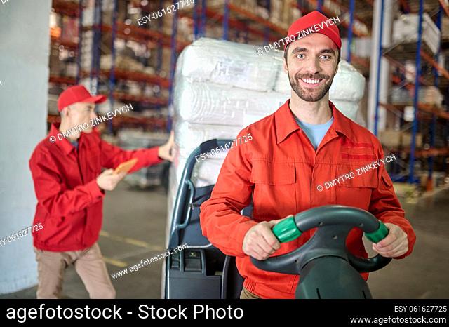 Smiling cheerful storehouse driver driving a forklift truck with cargo supervised by his serious colleague