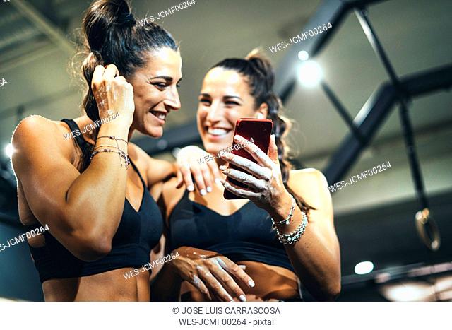 Smiling female twins in good shape using smartphone in a gym