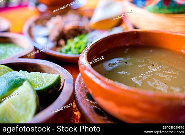 Close-up of clay bowls of lime slices, green sauce, and lamb broth with blurry chopped meat as background. Condiments and sliced vegetables above orange and red...