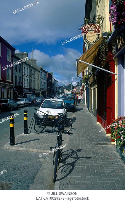 Shops/ houses/ buildings in High Street. Cars parked. Bicycles. People