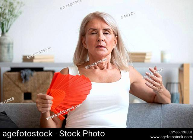 Tired mature woman suffering from heat at home, feeling uncomfortable, waving blue fan, sitting on couch, enjoying fresh air