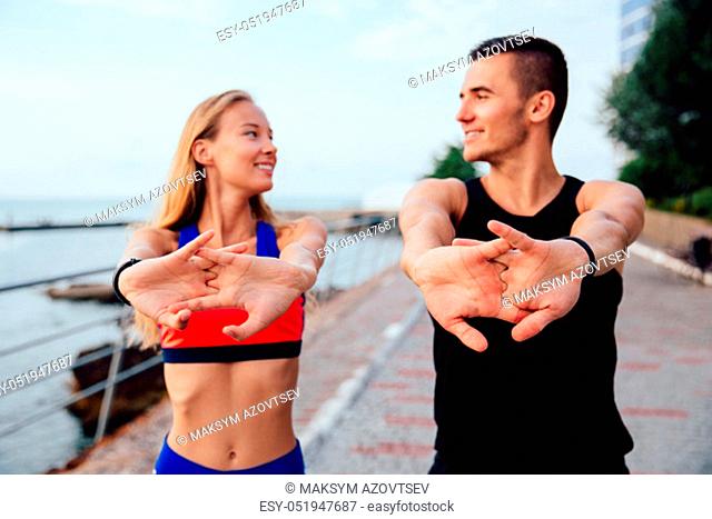 Happy man and smiling woman doing stretching exercises for arms during workout on the quay. Dressed in sportswear