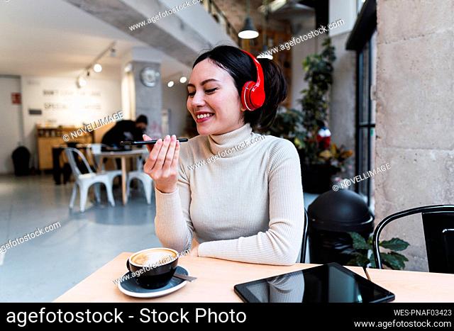 Smiling woman wearing headphones sending voicemail through smart phone at cafe