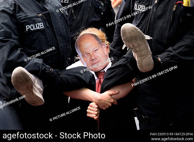 13 September 2023, Berlin: Police officers carry a participant wearing a mask with the likeness of Scholz off the street in front of the main train station...