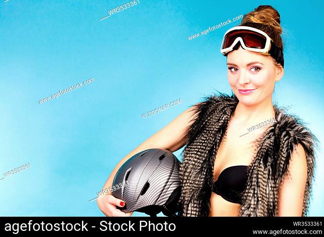 Woman sexy hot skier girl wearing black bra fur vest ski googles holding helmet. Winter sport activity. Beautiful seductive sportswoman on blue studio shot
