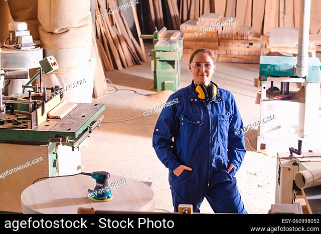 Portrait of young attractive girl in blue coverall, posing with hands folded on chest at warehouse interior. Professional female carpenter feeling proud doing...