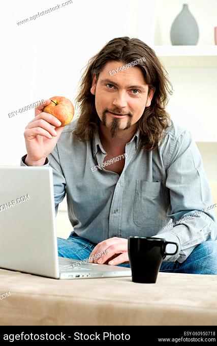 Casual man using laptop computer at home, sitting at couch, holding apple