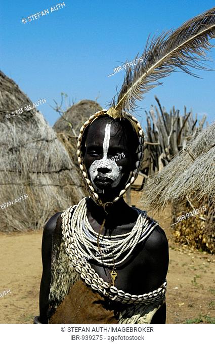Young Karo tribesman wearing feathers in a headband and necklaces of Kauri Shells standing between straw huts, portrait, Kolcho, South Omo Valley, Ethiopia