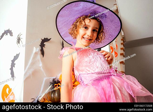 Portrait of adorable preschool girl with curly hair wearing fancy pink dress and purple witch hat posing at home. Staricase and walls with halloween decorations...