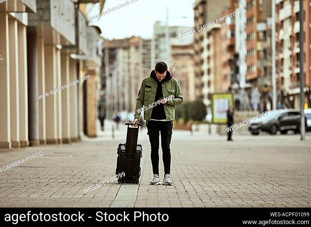 Young man with suitcase looking at mobile phone while standing on footpath in city