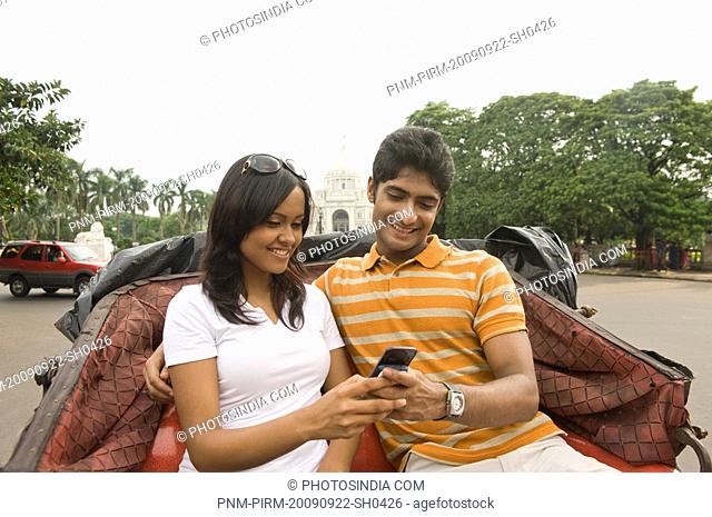 Couple sitting on a cart and using a mobile phone with a memorial in the background, Victoria Memorial, Kolkata, West Bengal, India