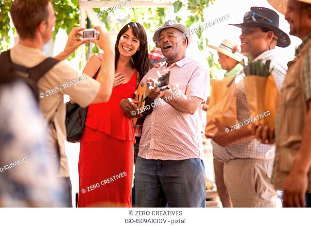 Tourist taking photograph of friend posing with market trader playing ukulele