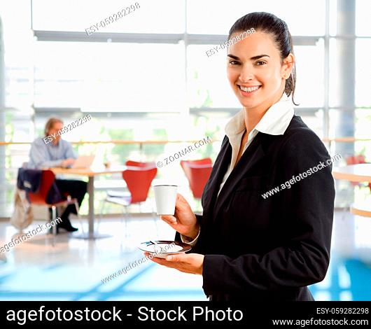 Happy young businesswoman standing in corporate building lobby, holding coffee cup, smiling