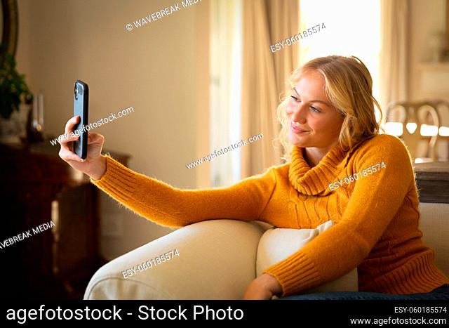 Smiling caucasian woman sitting on couch relaxing in living room taking smartphone selfie