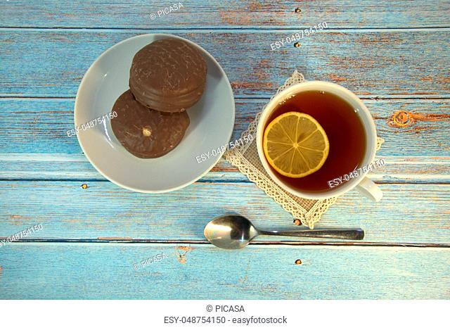 A cup of tea with lemon, a spoon and two chocolate cakes on a plate lie on a wooden table. Close-up