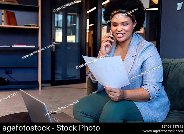 Smiling mid adult biracial businesswoman talking on smart phone while reading document in office