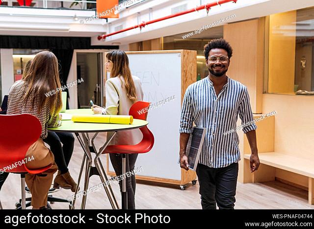 Smiling businessman holding solar panel in office with colleagues in background