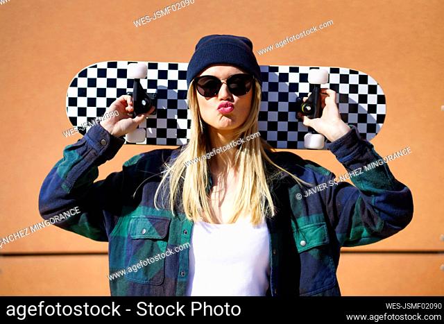 Blond woman puckering while carrying skateboard on shoulder in front of brown wall