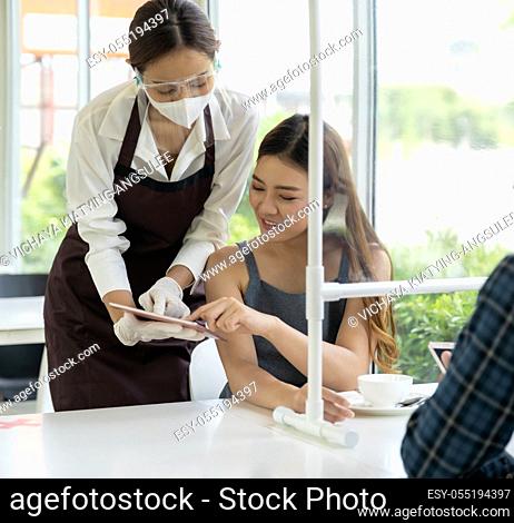 Asian waitress wear facemask and face shield using tablet to show restaurant electronic menu to reduce touching and recommend to customer