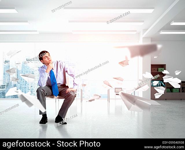 young businessman sits on an office chair. thoughtful and looking up. paper plane fly around