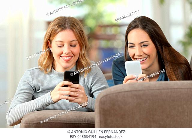 Front view of two happy roommates using two smart phones lying on a couch in the living room at home