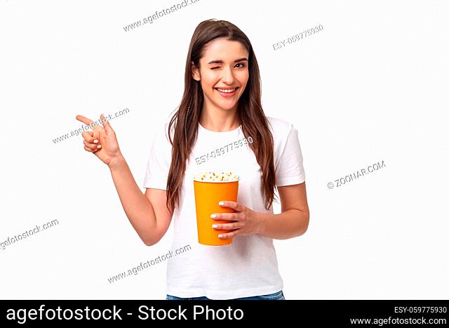 Entertainment, fun and holidays concept. Portrait of cheeky cute young girl in t-shirt, wink and smiling, pointing finger left inviting come and watch movie...