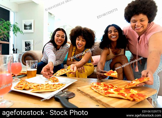 Excited biracial female friends picking pizza slices on table while spending leisure time at home