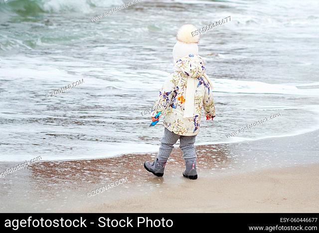 Cute little girl playing on the sandy beach. Happy child wearing warm floral print jacket, pom pom hat and scarf playing outdoors on fall, winter or spring day