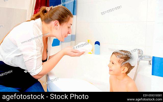 Happy smiling mother and little boy playing with soap foam in bath at home. Concept of child hygiene and health care at home